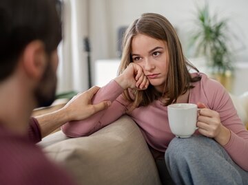 Eine junge Frau mit einer Tasse in der Hand sitzt einem Mann gegenüber auf der Couch und schaut unglücklich aus. | © GettyImages/Halfpoint Images