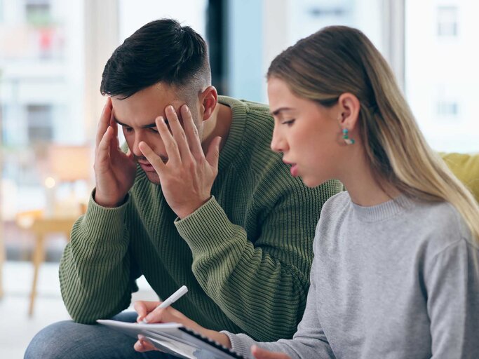Paar sitzt auf der Couch und diskutiert hitzig, während die Frau eine Liste schreibt. | © Adobe Stock/Charlize D/peopleimages.com