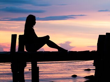 Frau sitzt allein auf einem Steg im Sonnenuntergang | © AdobeStock/photosky99
