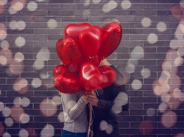 Paar steht vor einer Backsteinwand und küsst sich hinter vielen Herzluftballons. | © Adobe Stock/detailblick-foto