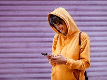 Frau mit gelbem Hoodie blickt auf ihr Handy vor lila Wand | © Getty Images/Westend61