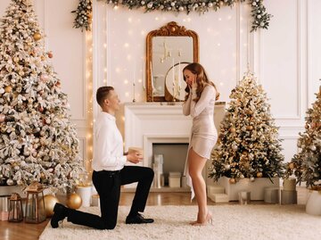 Mann macht Frau im Wohnzimmer mit Weihnachtsbaum einen Antrag | © Getty Images/Elizaveta Starkova