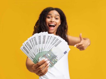 Aufgeregte afroamerikanische Frau hält mehrere Hunderteuro Scheine in der Hand und zeigt mit dem Finger auf Banknoten, sie steht vor einem gelbem Hintergrund. | © Adobe Stock/Prostock-studio