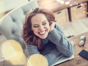 Junge glückliche Frau sitzt auf den Ellenbogen gestützt am Tisch und grinst breit. | © Adobe Stock/drubig-photo