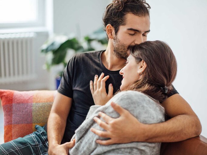 Junges Pärchen sitzt auf dem Sofa und Frau liegt in den Armen des Mannes.  | © Getty Images / Luis Alvarez