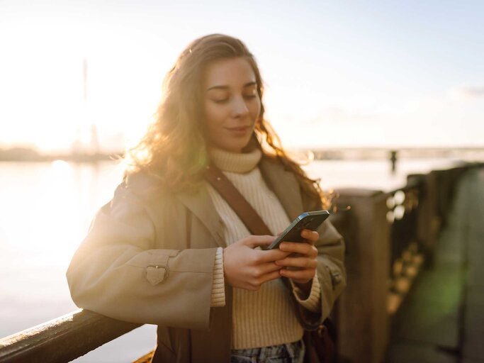 Frau steht im Sonnenuntergang im Herbst auf einem Steg und schreibt mit ihrem Handy eine Nachricht. | © Adobe Stock/maxbelchenko