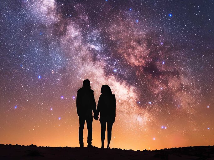 Ein Paar steht Hand in Hand vor der beeindruckenden Milchstraße und vielen Sternen, deren Licht den klaren Nachthimmel erhellt. | © Adobe Stock/Kateryna Kordubailo/Mit Ki generiert