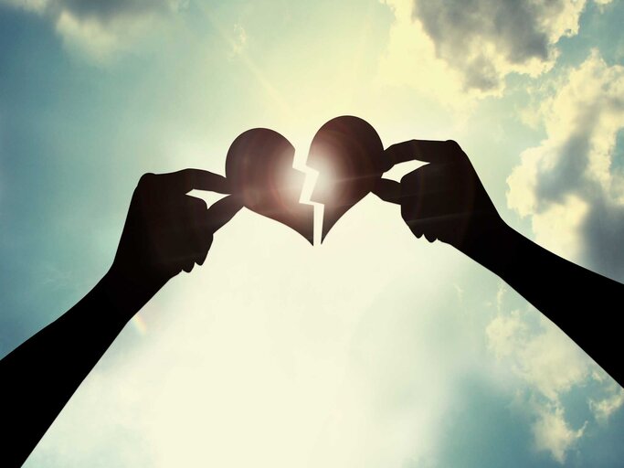 Zwei Hände halten ein zerrissenes Herz gegen die Sonne in den Himmel. | © Adobe Stock/2jenn
