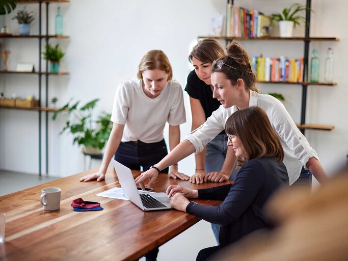 Eine Gruppe von Frauen steht an einem Tisch und schaut auf einen Laptop. | © Getty Images / Oliver Rossi