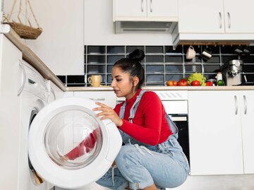 Frau mit Latzhose kniet vor einer Waschmaschine und gibt Kleidung hinein. | © Getty Images / jose carlos cerdeno martinez