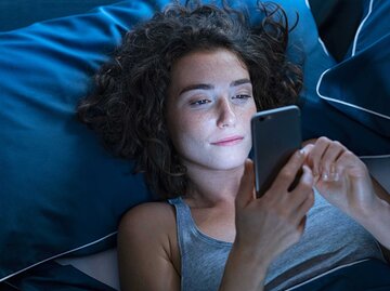 Frau liegt abends im Bett und schaut auf ihr Handy. | © Getty Images / Ridofranz
