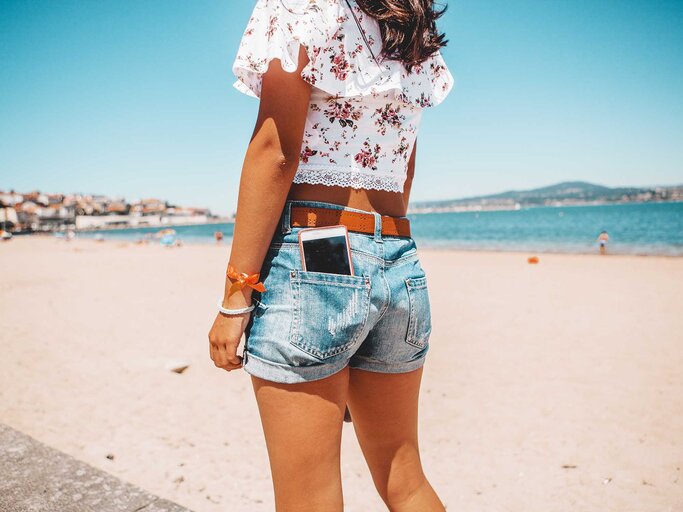 Frau mit kurzer Short und Bluse trägt ihr Handy in der hinteren Hosentasche und steht am Strand. | © Getty Images / Carol Yepes