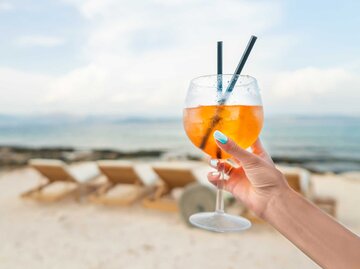 Die Hand einer Frau hält ein Glas mit einem Aperol Paloma am Strand in die Luft. | © Getty Images / repinanatoly