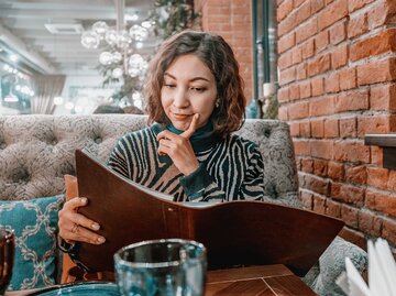 Junge junge Frau sitzt im Restaurant und schaut fragend in die Karte. | © Adobe Stock/EdNurg
