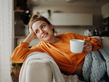 Eine junge Frau sitzt lächelnd auf dem Sofa mit einer Tasse in der Hand | © GettyImages/Halfpoint Images