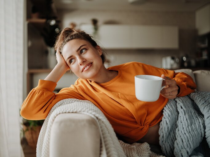 Eine junge Frau sitzt lächelnd auf dem Sofa mit einer Tasse in der Hand | © GettyImages/Halfpoint Images