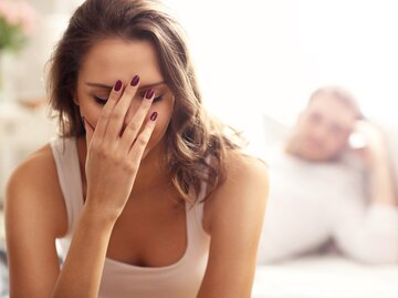 Junge Frau sitzt frustriert im Schlafzimmer. Mann sitzt im Hintergrund | © shutterstock/Kamil Macniak