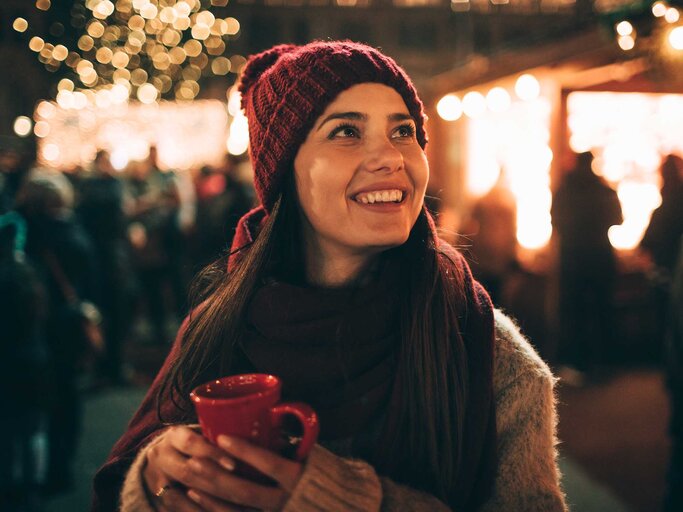 Frau auf dem Weihnachtsmarkt mit Lichtern im Hintergrund | © Getty Images/AleksandarNakic