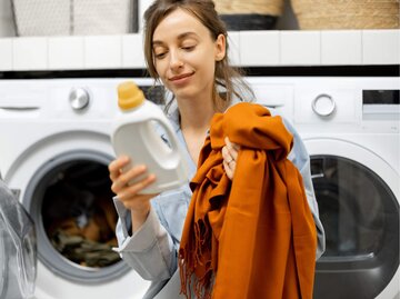 Frau mit Schal und Waschmittel vor Waschmaschine | © Getty Images/RossHelen