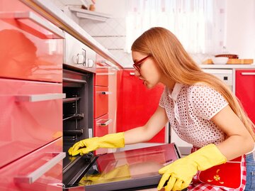 Junge Frau sitzt vor dem Ofen auf dem Boden und putzt das Backblech. | © Getty Images / stock_colors