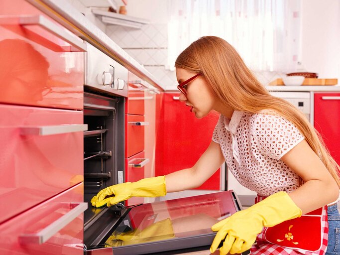 Junge Frau sitzt vor dem Ofen auf dem Boden und putzt das Backblech. | © Getty Images / stock_colors
