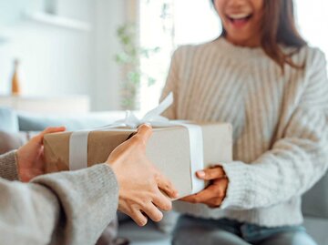 Eine lachende Frau in beigem Strickpullover bekommt ein verpacktes Weihnachtsgeschenk mit weißer Schleife von einer Person im grauen Pullover. | © Adobe Stock/oatawa