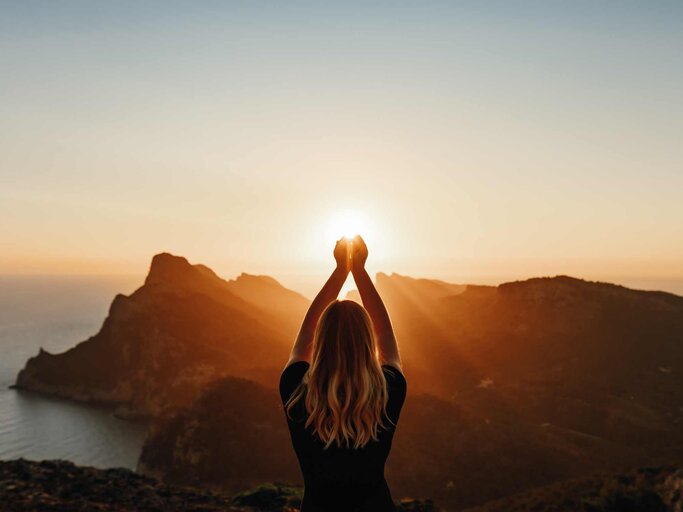 Frau steht auf Berg vor dem Sonnenuntergang mit den Händen in der Luft | © Getty Images/DianaHirsch