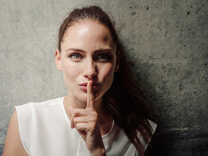 Junge Frau mit Finger auf Lippen vor einer Wand | © Getty Images/Portra