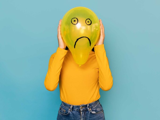 Frau hält sich gelben Luftballon mit traurigem Smiley vor das Gesicht | © Getty Images/Xavier Lorenzo