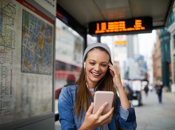 Eine lächelnde junge Frau mit einer weißen Strickmütze und einem blauen Jeanshemd steht an einer Bushaltestelle. Sie trägt Ohrhörer und schaut auf ihr Smartphone.  | © Adobe Stock/Geber86