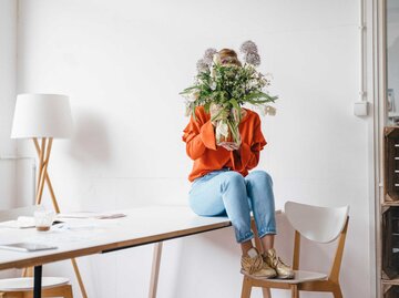 Frau sitzt auf einem Tisch und hält sich eine Blumenvase vor ihr Gesicht. | © Getty Images/Westend61