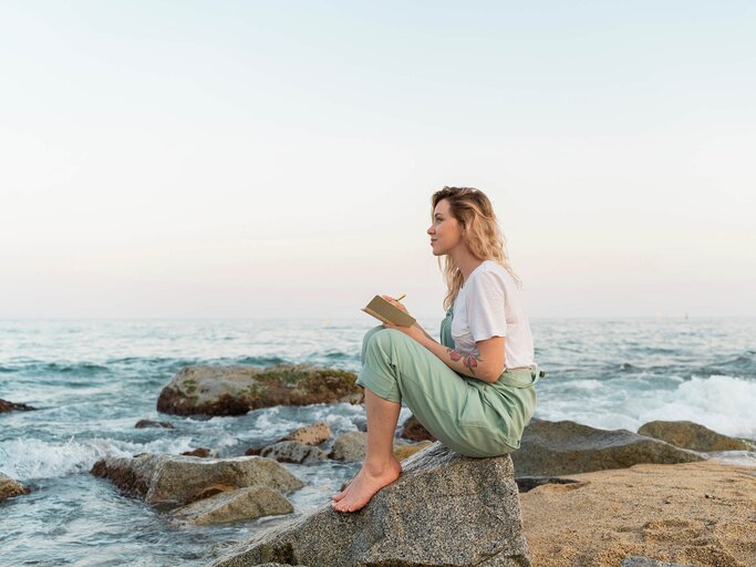 Frau mit Tagebuch auf dem Schoß sitzt auf einem Felsen am Meer | © Getty Images/Westend61