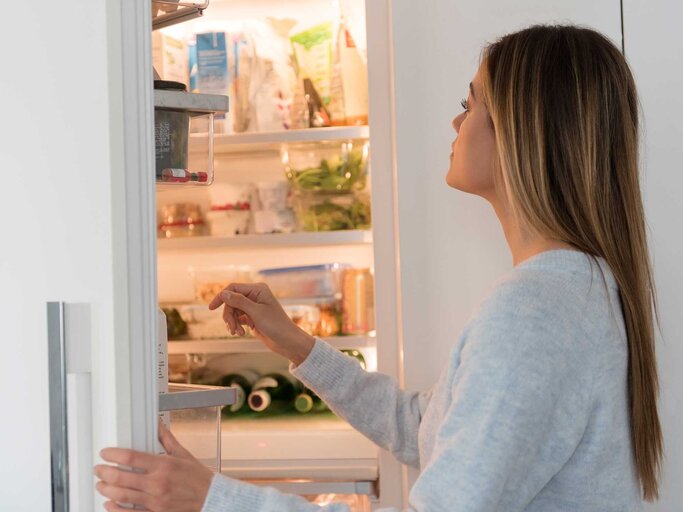 Junge Frau steht vor dem geöffneten Kühlschrank und schaut hinein. | © Getty Images / Jamie Grill