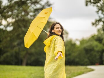 Eine Frau im Regenmantel und mit Regenschirm streckt die Hand nach hinten aus | © GettyImages/Eleganza