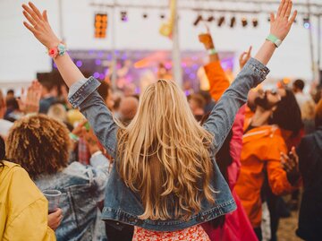 Frau steht auf einem Festival mit dem Rücken zur Kamera und streckt ihre Hände in die Luft. | © Getty Images / SolStock