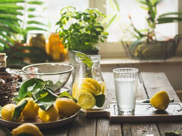 Glaskrug mit frischer Zitrone und Glas Limonade auf hölzernem Küchentisch mit Zutaten am Fenster. | © Getty Images/VICUSCHKA