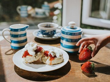 Hand greift nach einer Erdbeere, darunter ein Holztisch mit einem Teller Scones mit Schlagsahne und frischen Erdbeeren | © Getty Images/Catherine Falls Commercial