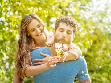 Ein Mann pustet an einer Pusteblume und eine Frau lächelt wegen ihrer Frühlingsgefühle | © GettyImages/Arne Trautmann/EyeEm