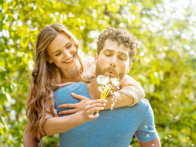 Ein Mann pustet an einer Pusteblume und eine Frau lächelt wegen ihrer Frühlingsgefühle | © GettyImages/Arne Trautmann/EyeEm