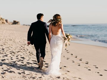 Zwei Personen in Hochzeits-Outfits rennen Hand in Hand am Strand | © Getty Images/Westend61