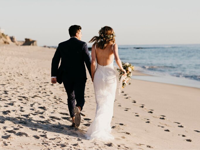 Zwei Personen in Hochzeits-Outfits rennen Hand in Hand am Strand | © Getty Images/Westend61
