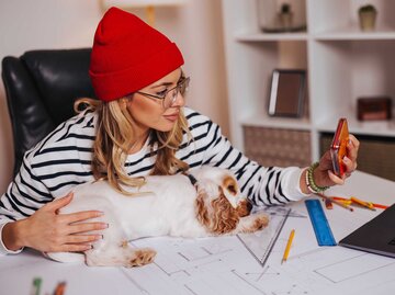 Frau mit roter Mütze sitzt am Arbeitsplatz und macht ein Selfie von sich und ihrem Hund. | © Getty Images / bojanstory