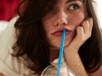 Junge Frau mit Sommersprossen trinkt gelangweilt aus einem Strohalm | © Getty Images/ Westend61