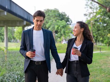 Junges Business-Paar läuft mit Kaffee in der Hand Händchenhaltend durch den Park. | © Getty Images / Jackyenjoyphotography