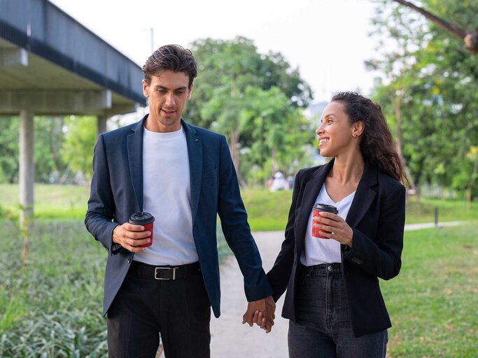 Junges Business-Paar läuft mit Kaffee in der Hand Händchenhaltend durch den Park. | © Getty Images / Jackyenjoyphotography