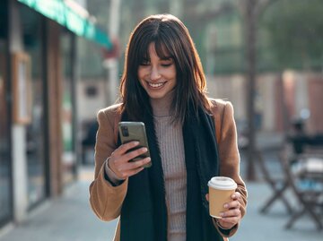 Junge Frau läuft auf der Straße mit einem Kaffeebescher in der Hand und schaut in ihr Handy. | © Adobe Stock/nenetus
