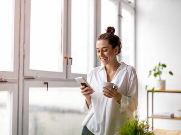 Frau steht im Zimmer und schaut auf ihr Handy. | © Adobe Stock/pikselstock