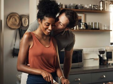 Mann küsst seine Partnerin auf die Wange während sie kocht | © Adobe Stock/Allistair/peopleimages.com