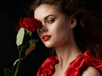 Frau mit langen, braunen Haaren trägt rotes Kleid und hält rote Rose in der Hand | © AdobeStock/SHOTPRIME STUDIO