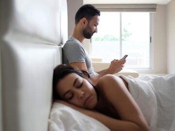 Paar ist im Bett, Frau schläft und Mann guckt aufs Handy | © Getty Images/Diego Cervo / EyeEm